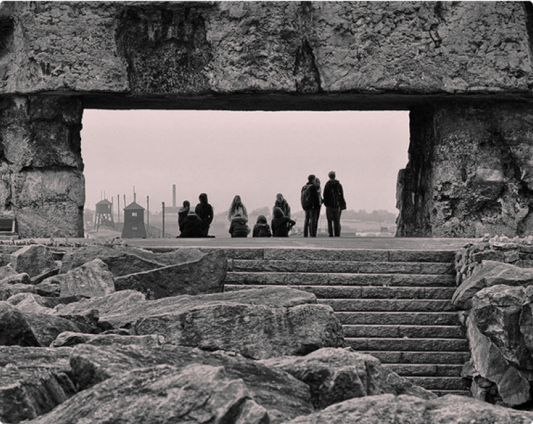 Zdjęcie pokazuje kamienie i kamienne schody, na których stoi grupa ludzi. W tle ogrodzenie, wieżyczki i komin