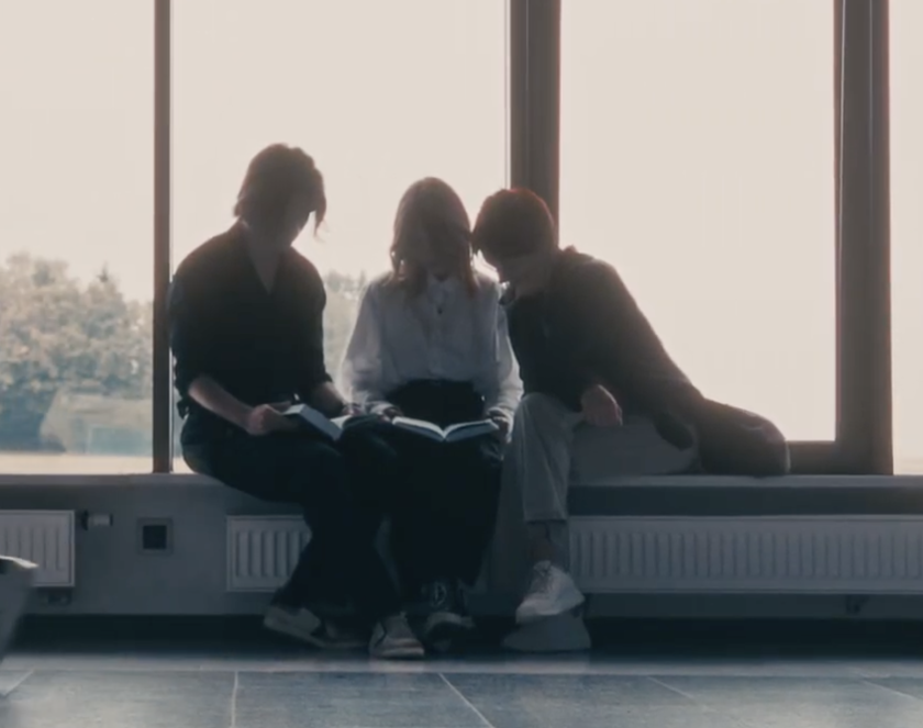 Młody mężczyzna i dwie kobiety czytają książkę na tle dużych okien.