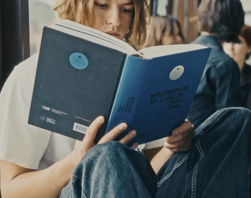 Młody mężczyzna o dłuższych włosach czytający książkę z fioletową okładką. 