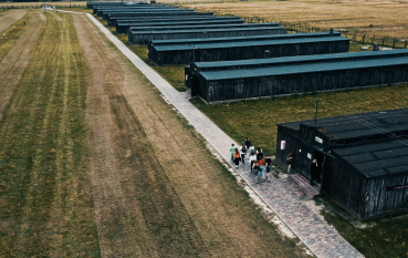 Widok z lotu ptaka, rząd drewnianych baraków więźniarskich, na drodze na wysokości baraku pierwszego a drugiego grupa zwiedzających.