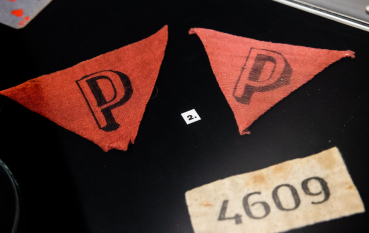 Dwa trójkątne czerwone winkle więźniarskie z czarną literą P stylizowaną na trójwymiarową oraz numer obozowy 4609 wykonany na prostokątnym skrawku płótna