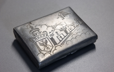 Metalowa papierośnica z ręcznie wykonanym grawerem przedstawiającym wieżę z orłem oraz trzy baraki więźniarskie.