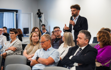 Mężczyzna w średnim wieku w okularach i z brodą stoi i przemawia do mikrofonu.