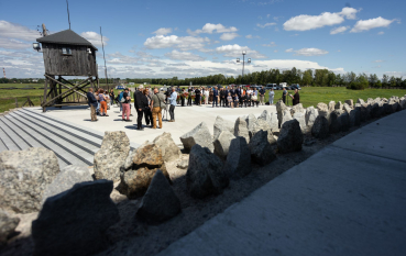 Grupa osób zebrana pod Pomnikiem Mauzoleum, na pierwszym planie rząd ostrych kamieni, na drugim wieża wartownicza