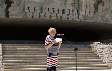 Starsza kobieta ubrana w sukienkę w kolorowe poprzeczna pasy stoi pod napisem wyrytym na fryzie Mauzoleum: Los nasz dla Was przestrogą 