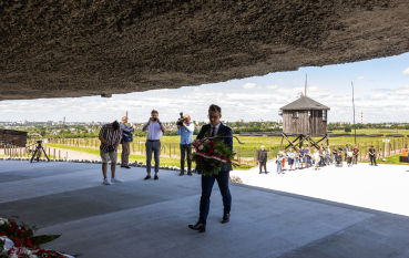 Mężczyzna w garniturze niesie wieniec na platformie pomnika Mauzoleum, na drugim planie fotoreporterzy