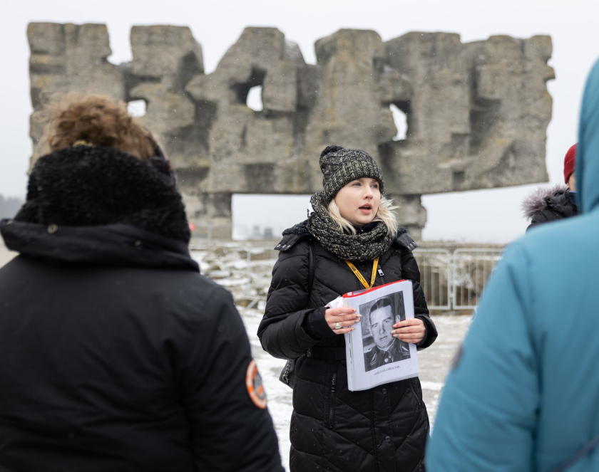 Kobieta ubrana w zimową kurtkę i czapkę na tle Pomnika Bramy trzyma w rękach czarno białe zdjęcie niemieckiego oficera. 