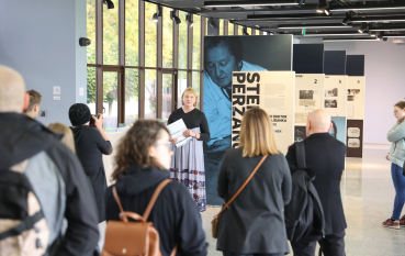 Kobieta w długiej sukni na tle drukowanej wystawy na terenie obiektu, otoczona kilkoma osobami. 