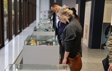 Grupa osób oglądająca przedmioty w przeszklonych gablotach, na pierwszym planie kobieta o blond włosach ubrana w melanżowy golf.