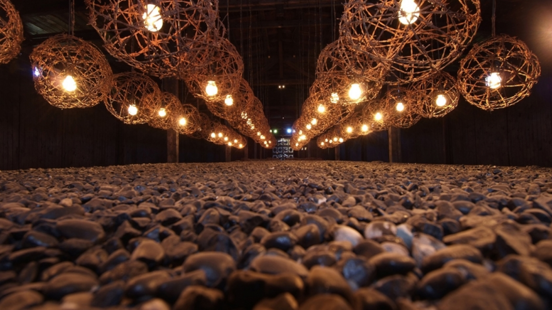 Zdjęcie instalacji artystycznej. W czerech rzędach zawieszone kule z drutu kolczastego z żarówkami w środku, poniżej usypane kamienie.