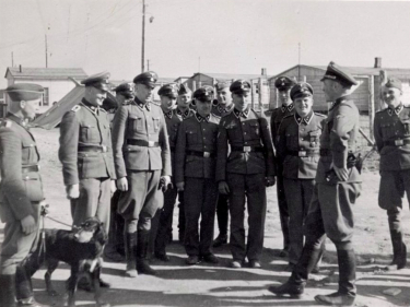 Grupa kilkunastu mężczyzn w niemieckich mundurach. Jeden z nich trzyma na smyczy psa. W tle baraki