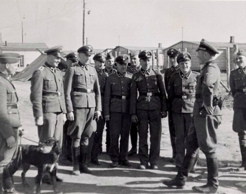 Grupa kilkunastu mężczyzn w niemieckich mundurach. Jeden z nich trzyma na smyczy psa. W tle baraki
