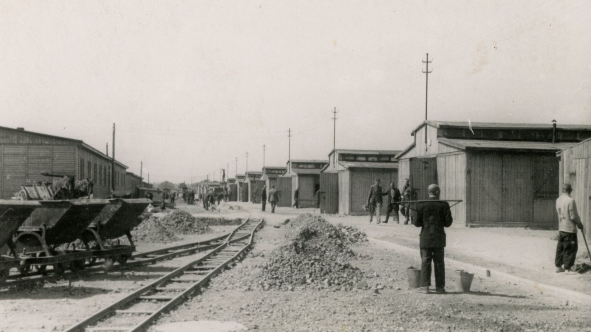 Czarno-białe zdjęcie, droga po lewej stronie tory kolejki i wagonik, po prawej pracujący mężczyźni. 