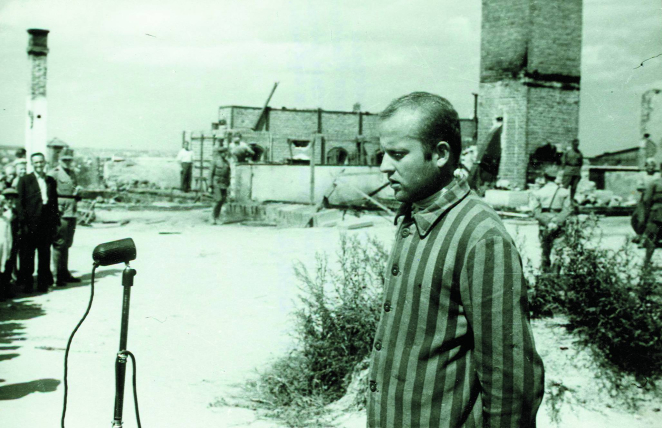 Mężczyzna w pasiaku stoi przed mikrofonem i mówi. Z nim murowany piec i wysoki komin. Dookoła pojedynczy ludzie