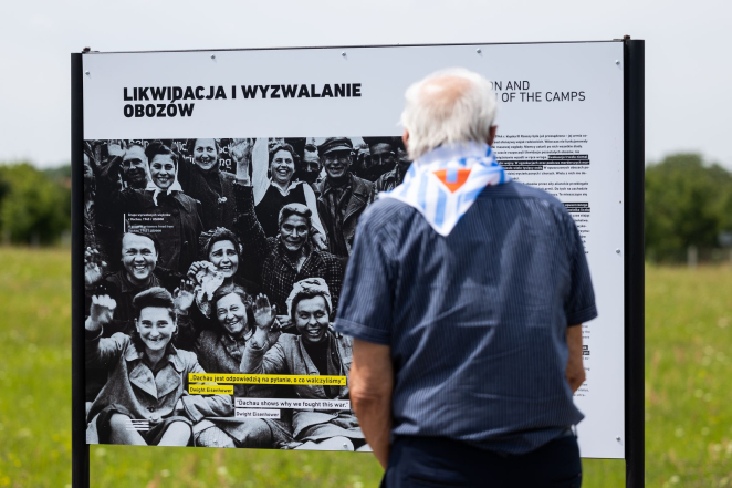 Starszy mężczyzna stojący tyłem, na szyi chusta w biało niebieskie pasy i czerwony trójkąt z literą P, na drugim planie plansza z wystawy plenerowej zatytułowana: Likwidacja i wyzwalanie obozów. 