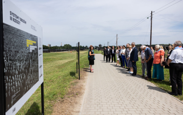 Grupa osób ustawionych rzędami, kilka osób w średnim wieku ubranych w eleganckie ubrania oraz grupa starszych osób noszących chusty w biało-niebieskie pasy, na pierwszym planie plansza wstawy: system obozów III Rzeszy. 