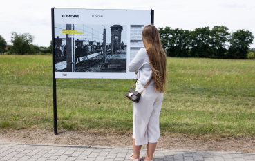 Kobieta ubrana w jasne ubrania o długim blond włosach przed tablicą wystawy plenerowej; napis na tablicy: KL Dachau 