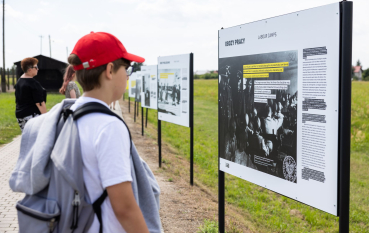Nastoletni chłopiec w czerwonej kaszkietówce stoi przed plansza\ą wystawy plenerowej; napis na planszy: Obozy pracy 