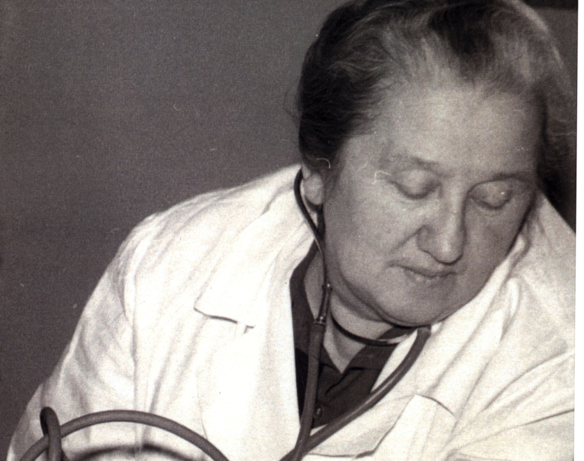 Czarno-biały portret lekarki mierzącej ciśnienie pacjentowi , kobieta w starszym wieku ubrana w lekarski kitel, na szyi stetoskop, obok ręka pacjenta i ciśnieniomierz. 