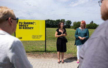 Dwie kobiety, jedna ubrana w czarną obcisłą sukienkę z dekoltem caro, druga w zieloną obszerną tunikę, obok żółta plansza wystawy, napis: Tu Śmierć Obozy III Rzeszy, Death is Here, Camps of the Third Reich. 