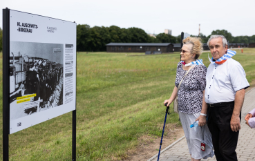 Starsza kobieta wspierająca się o laskę i starszy mężczyzna, oboje w chustach w niebiesko-białe pasy, obok plansza z wystawą napis: KL Auschwitz-Birkenau 