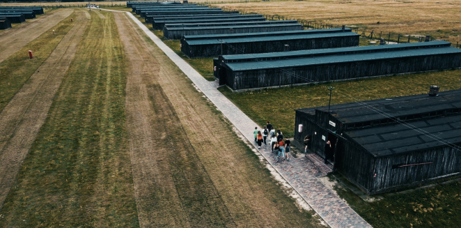 Zdjęcie z drona, rząd baraków więźniarskich obok jednego z nich idąca ścieżką grupa osób 