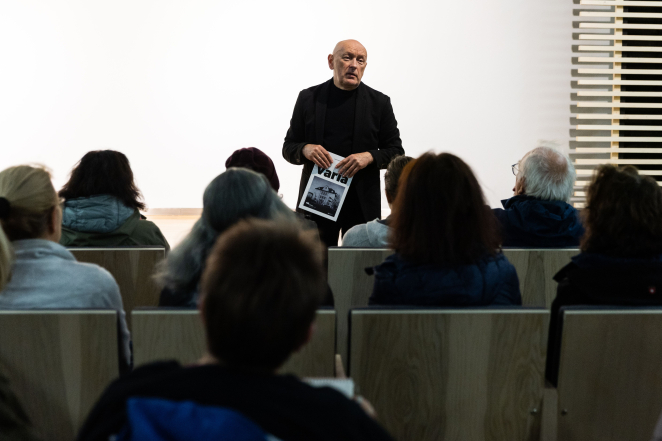 Łysy mężczyzna w średnim wieku ubrany na czarno stoi naprzeciw grupy osób siedzących na sali audytoryjnej, w rękach trzyma magazyn Varia 
