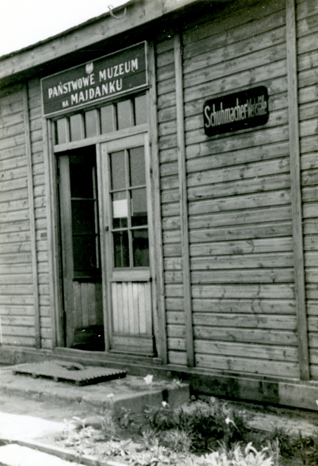 Wejście do drewnianego baraku, otwarta jedna część drzwi, nad nimi napis Państwowe Muzeum na Majdanku oraz polskie godło, po prawej stronie napis w języku niemieckim.
