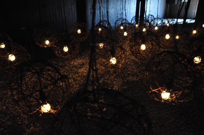 Kilkanaście sfer z drutu kolczastego z umieszczonymi po środku żarówkami, dookoła ciemne pomieszczenie