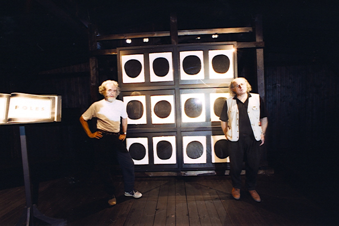 Dwaj mężczyźni o siwych długich do ramion włosach, mężczyzna po lewej ubrany w biały t-shirt, mężczyzna po prawej ubrany w czarny t-shirt i białą kamizelkę, w tle instalacja artystyczna: 12 białych prostokątnych kart z czarnym kołem pośrodku.