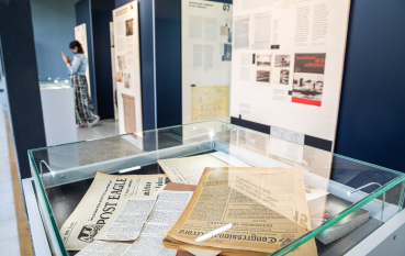 Na pierwszym planie szklana gablota z wycinkami z gazet, w tle postać kobiety