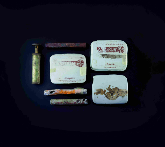 Trzy prostokątne metalowe pudełka po lekach: ASPIRIN BAYER, dwie kolorowe szklane fiolki, dwa metalowe podłużne przedmioty. 