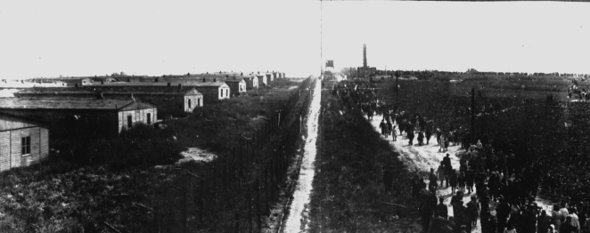 Czarno białe zdjęcie, teren obozu koncentracyjnego, na środku droga, po prawej stronie rząd drewnianych baraków po lewej kilkadziesiąt osób.