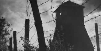 Czarno-białe zdjęcie. Poprzez druty kolczaste widać z prawej strony wieżę strażniczą. Z prawej strony liście 