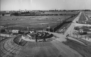 Czarno-białe zdjęcie, kopuła Mauzoleum widziana z lotu ptaka, dwóch mężczyzn pracuje przy budowie okulusa, w tle teren byłego obozu koncentracyjnego. 
