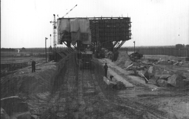 Czarno-białe zdjęcie, Pomnik-Brama pokryty rusztowaniem oraz płachtami zabezpieczającymi, na pierwszym planie droga dojazdowa do budowy.