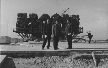 Trzech mężczyzn ubranych w garnitury na tle Pomnika-Bramy, w tle dźwig. 