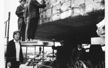 Kilku mężczyzn przy fryzie Mauzoleum, mężczyzna na pierwszym planie ubrany w białą bluzkę i czarną marynarkę, mężczyzna na drugim planie trzymający dłuto, wykuwający relief ubrany w strój roboczy. 