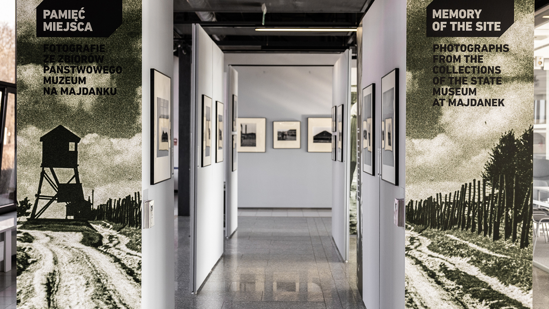 Widok wystawy, korytarz stworzony przez ścianki wystawiennicze po obu stronach, na których zawieszone są fotografie. Na pierwszym planie po lewej i prawej stronie tablice tytułowe wystawy.  Na zdjęciu z lewej wieżyczka wartownicza, z prawej ogrodzenie z drutu kolczastego. 