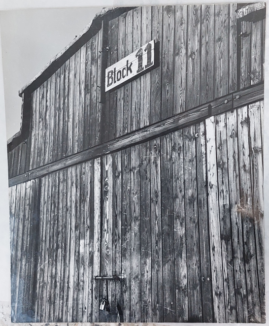 Czarno-białe zdjęcie, drewniana ściana baraku z drzwiami. W górnej części tabliczka z napisem: Block 11
