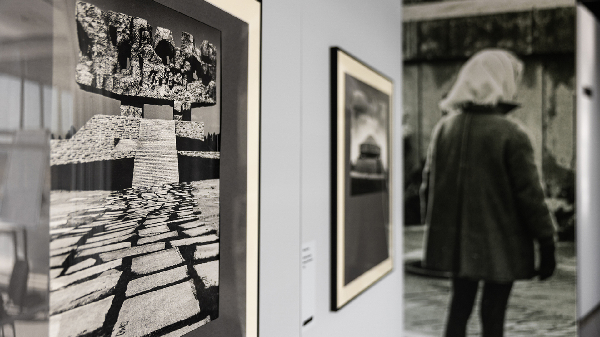 Widok wystawy, z lewej oprawione w ramę zdjęcie pomnika-bramy na Majdanku, obok zdjęcie mauzoleum, w tle nie zdjęcie dziewczynki obróconej tyłem do widza, w chustce na głowie i płaszczu.