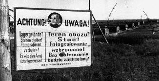 Czarno-białe zdjęcie tablicy w języku polskim i niemieckim: Achtung, Uwaga. Pośrodku grafika czaszki ze skrzyżowanymi kośćmi piszczelowymi. 