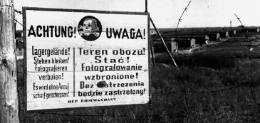 Czarno-białe zdjęcie tablicy w języku polskim i niemieckim: Achtung, Uwaga. Pośrodku grafika czaszki ze skrzyżowanymi kośćmi piszczelowymi. 