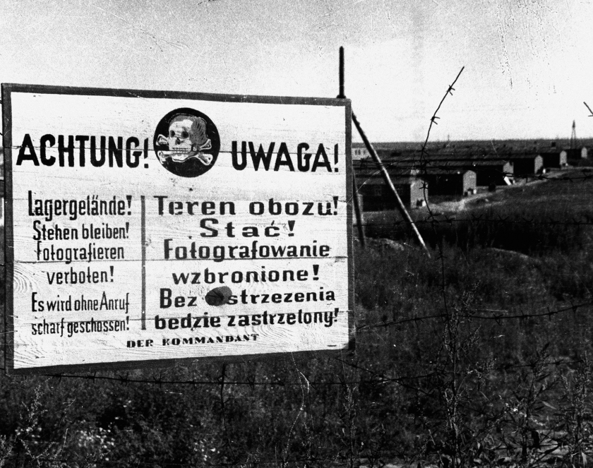 Czarno-białe zdjęcie tablicy w języku polskim i niemieckim: Achtung, Uwaga. Pośrodku grafika czaszki ze skrzyżowanymi kośćmi piszczelowymi. 