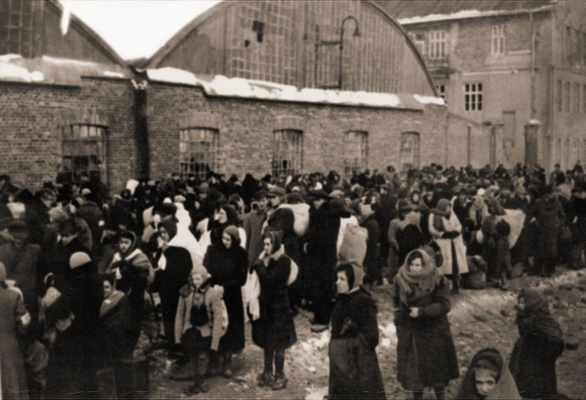 Czarno-białe zdjęcie, duża grupa osób z tobołkami, w tle zabudowania, hangary.