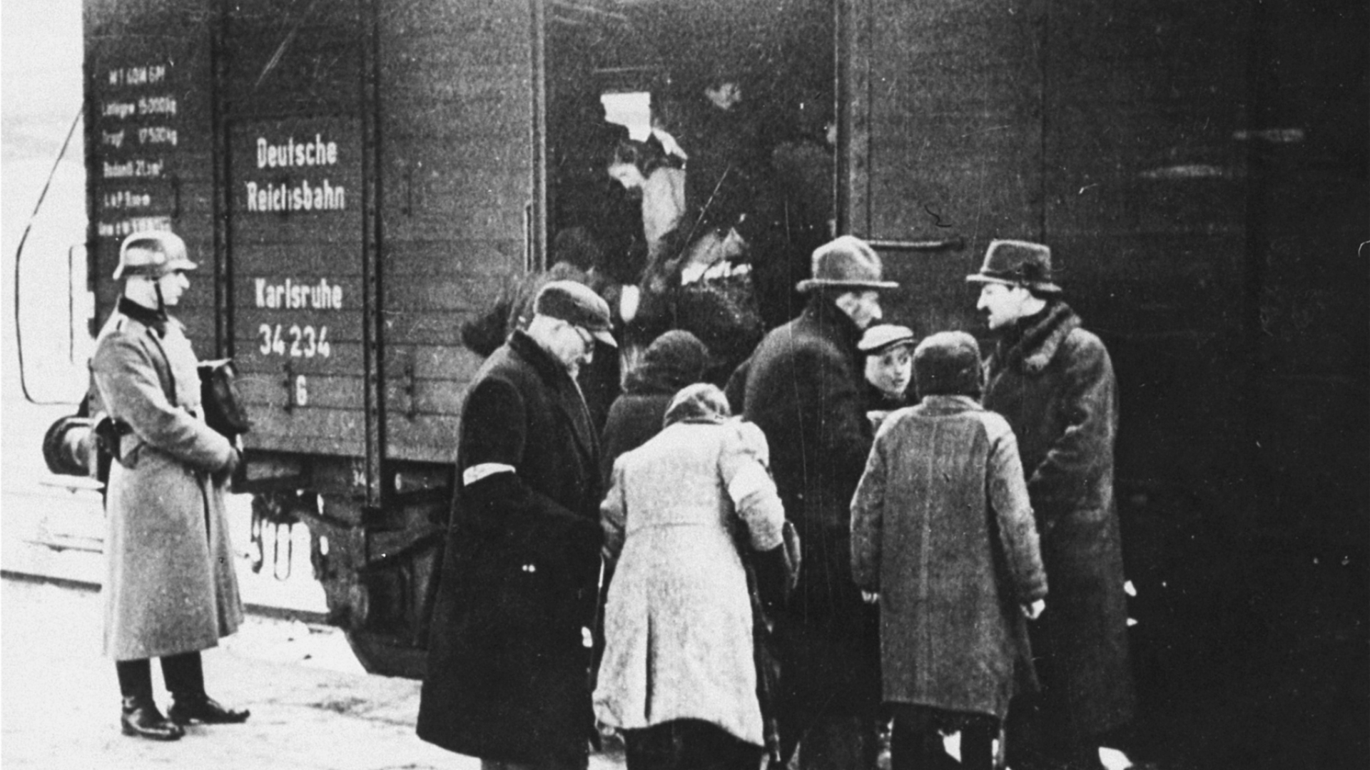 Zdjęcie czarno-białe, historyczne. Ludzie stojący w grupie przy wagonie pociągu. Mężczyzna w ciemnym płaczu z opaską na ramieniu. Po lewej stronie soi żołnierz w płaszczu i hełmie. W otartych drzwiach wagonu widać innych ludzi.  