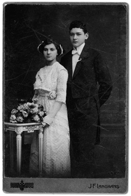 Czarno-białe zdjęcie ślubne: kobieta ubrana w białą koronkową suknię ślubną o upiętych wysoko czarnych włosach, mężczyzna ubrany w czarny garnitur, białą koszulę z biała muszką; przed parą młodą stolik i ułożony na nim bukiet jasnych kwiatów.