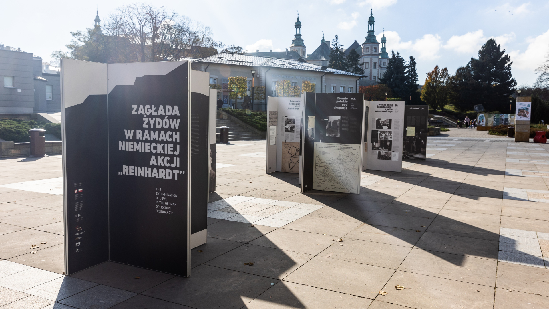 Zdjęcie współczesne, wystawa plenerowa stoi na placu. W tle zabudowania. 