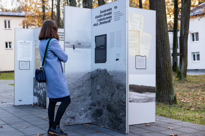Kobieta w prostym płaszczu do kolan oglądająca wystawę plenerową, na drugim planie kilka drzew i zabudowań.