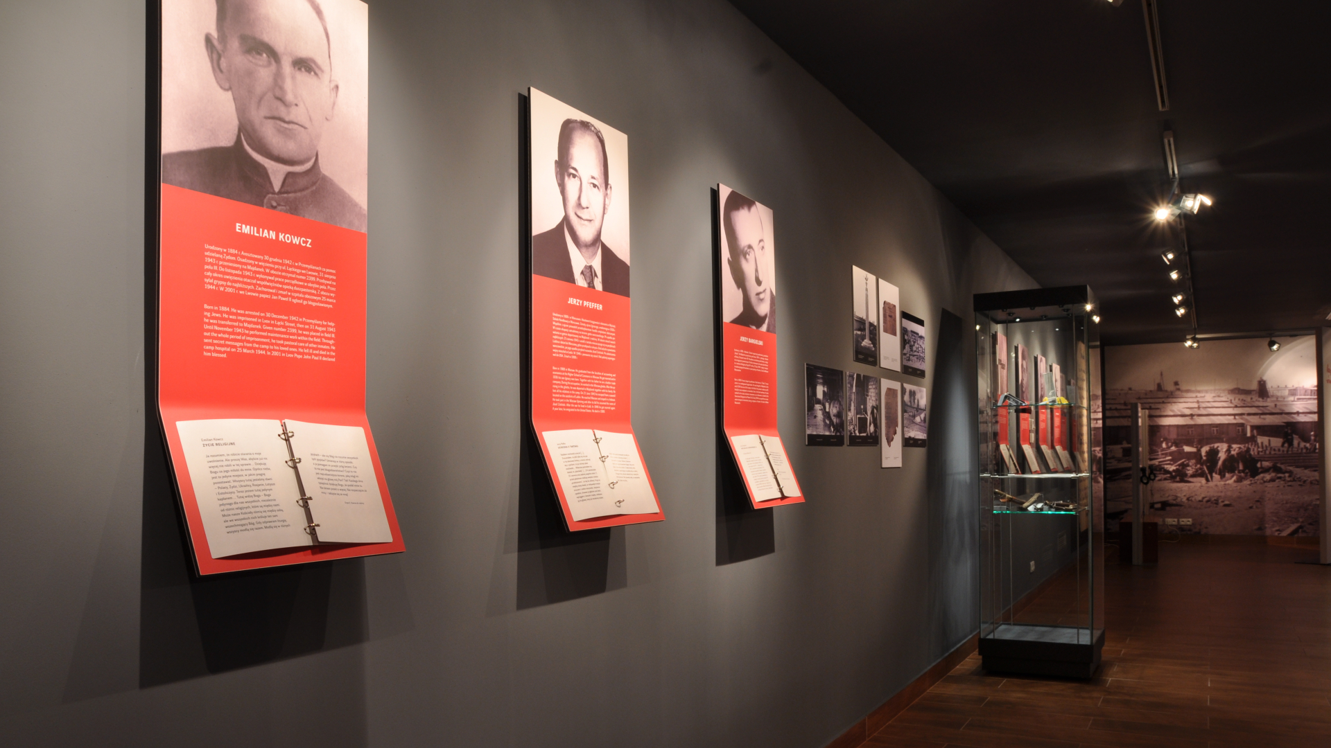 Widok na fragment wystawy. Na ścianie wiszą portrety więźniów, poniżej na tablicach tekst z biogramem. W oddali gablota.